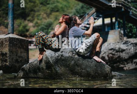 Two friends sit back-to-back on a sunlit rock by the river, meditating with hands clasped in prayer Stock Photo