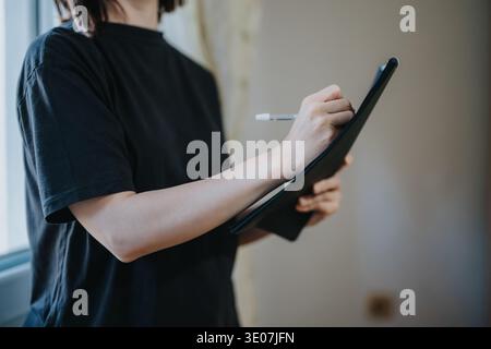 Woman writing on clipboard holding a pen and taking notes by a window in a casual shirt Stock Photo