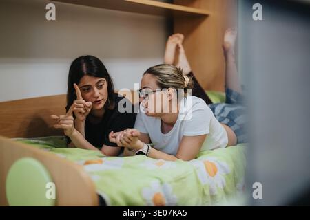Two women friends talking and sharing a conversation while lying on a bed in a bedroom Stock Photo
