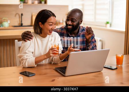 A joyful couple spends time together online, reacting happily to content on a laptop while enjoying drinks in a comfortable kitchen environment. Stock Photo