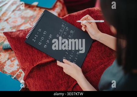 Student solving math problems in a black spiral notebook while studying at home on a red cushion Stock Photo