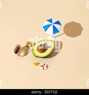 Funny avocado character relaxing on beach with beach umbrella and mini vacation accessories on sand color background. Summer concept. Stock Photo