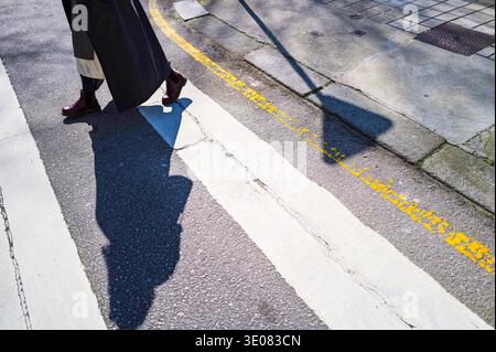 Person crossing street at a pedestrian crossing on a sunny day with shadows, including a “Yield” sign shadow Stock Photo