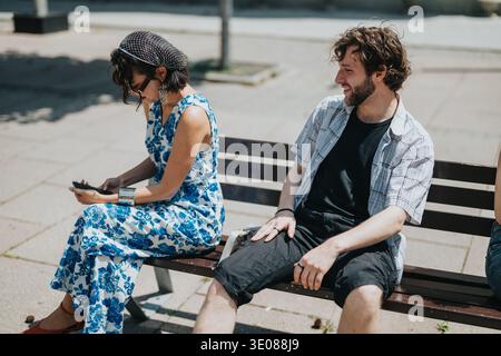 Two friends sit on a park bench, woman in blue floral dress using phone while man chats nearby Stock Photo