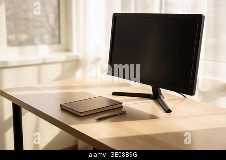 A clean wooden desk features a black computer monitor and a closed notebook with a pen, illuminated by soft sunlight streaming through a window with s Stock Photo