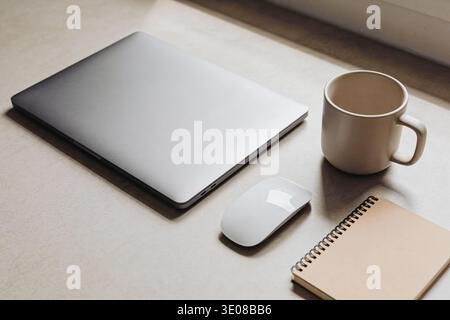A clean and organized desk setup featuring a closed laptop, a white wireless mouse, a spiral-bound notebook, and a ceramic mug, bathed in soft natural Stock Photo