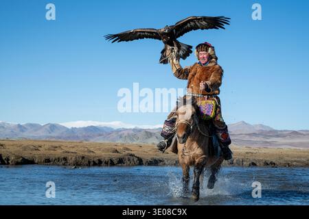 Mongolian eagle hunter (berkutchi) riding a horse in a river with a blindfolded golden eagle, Altai Mountains, Bayan-Olgii, Western Mongolia, Mongolia Stock Photo