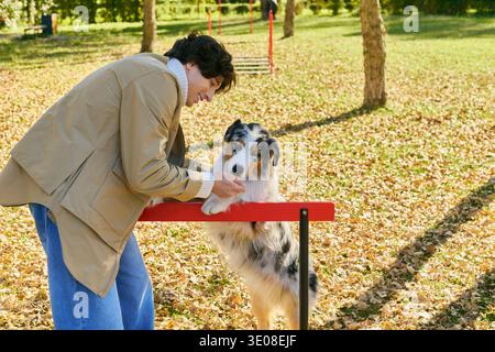 A young man with curly hair smiles with his Australian Shepherd among fallen leaves. Stock Photo