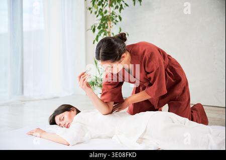 Masseuse making thai yoga massage. Woman therapist doing traditional massage treatment for female patient using elbow. Client lying on mat, meditating, enjoying hand massage in calm atmosphere. Stock Photo