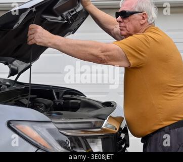 Senior man opens hood on vehicle to check engine. He wears sunglasses and a yellow shirt. His hands grip tools near the open hood. Vehicle is parked i Stock Photo