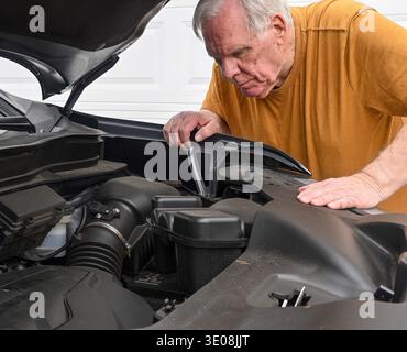 An older man in a yellow shirt works on a car engine. His hand holds a tool, the other steadies the hood. He concentrates intently, brow furrowed. Pra Stock Photo