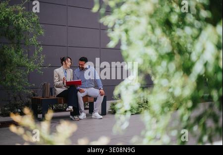 Business collaboration between two colleagues discussing documents on a bench outside an office building Stock Photo