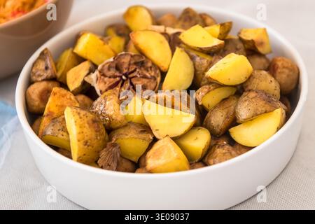 A white serving bowl filled with golden-brown roasted potato wedges. The dish is seasoned with herbs and features a whole head of roasted garlic in th Stock Photo
