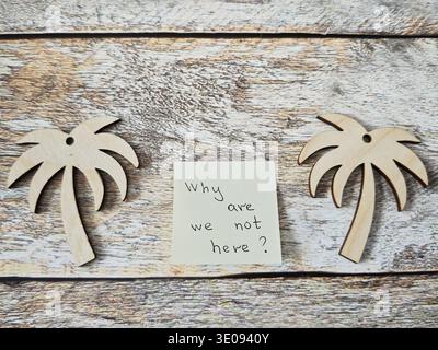 Two wooden palm tree shapes sit on a wood surface beside a sticky note that asks why the viewer is not on vacation. This reflects on travel savings an Stock Photo