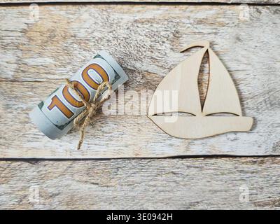 Rolled banknotes shown next to a wooden sailboat on a rustic surface, representing travel savings and budgeting for future vacations and holidays. Stock Photo