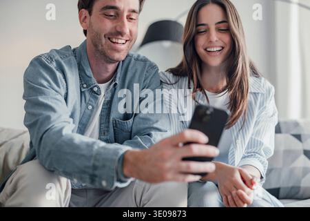 Happy millennial couple sit relax on couch in living room watching video on cellphone together, smiling young husband and wife rest on sofa at home br Stock Photo