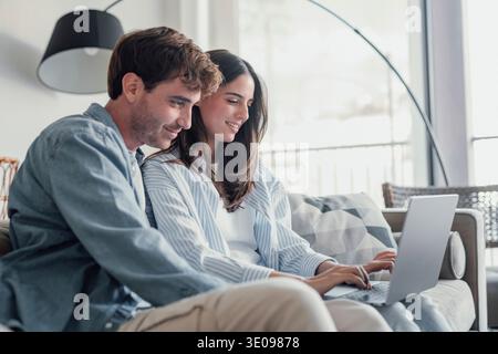 Happy millennial couple sit relax on couch in living room watching video on laptop together, smiling young husband and wife rest on sofa at home brows Stock Photo