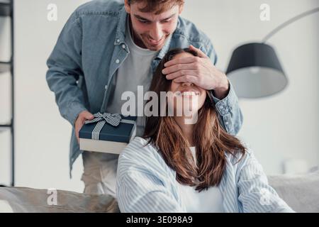 Excited young caucasian man giving surprise gift to excited girlfriend, standing back, putting hand on eyes, laughing. Joyful millennial couple laughi Stock Photo