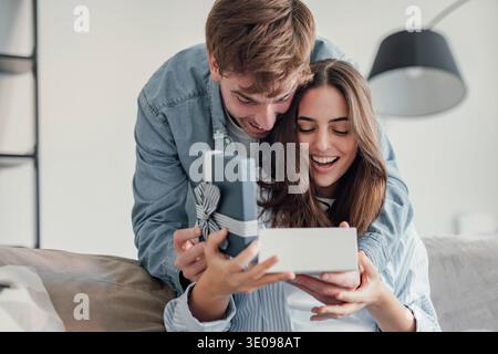 Excited young caucasian man giving surprise gift to excited girlfriend, standing back, putting hand on eyes, laughing. Joyful millennial couple laughi Stock Photo