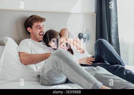 A young couple lies in bed, laughing and playfully teasing each other. Their smiles and playful touches show joy, closeness, and love, sharing a fun, Stock Photo