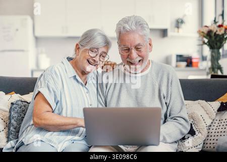 Pretty elderly 70s grey-haired couple resting on couch in living room hold on lap laptop watching movie smiling enjoy free time, older generation and Stock Photo