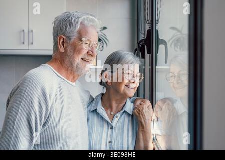 Happy bonding loving middle aged senior retired couple standing near window, looking in distance, recollecting good memories or planning common future Stock Photo