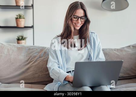 A young woman sits on the sofa wearing glasses, focused on her smartphone. She works, browses, and shops online at home, reflecting concentration, pro Stock Photo