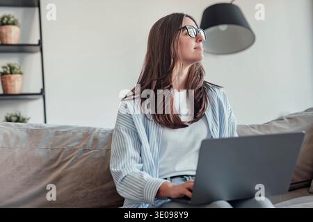 A young woman sits on the sofa wearing glasses, looking thoughtfully outside. She reflects on work, browsing, and lifestyle, showing concentration, co Stock Photo