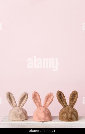 Three knitted bunny hats in beige, pink, and brown displayed on a white surface with a soft pink background creating a playful atmosphere Stock Photo