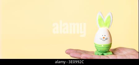 A hand holds a decorative Easter egg designed with bunny ears and a smiling face, set against a soft yellow background Stock Photo