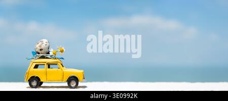 A yellow toy car is parked with quelle egg  on its roof, set against a bright blue sky and ocean view, creating a playful easter travel scene Stock Photo