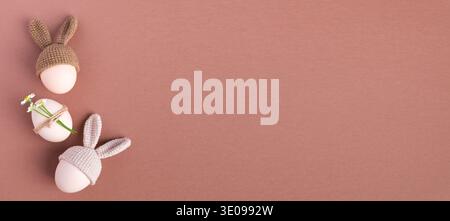 Three decorative Easter eggs wearing knitted bunny hats and adorned with flowers, arranged on a soft pink background for a festive spring theme Stock Photo
