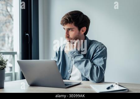 Thoughtful businessman touching chin, pondering ideas or strategy, sitting at wooden work desk with laptop, freelancer working on online project, stud Stock Photo