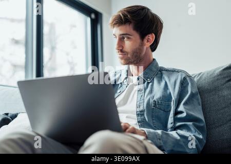 Dreamy pensive freelance man thinking over work project ideas on home office couch, using laptop, looking at window away, smiling at good thoughts, fu Stock Photo