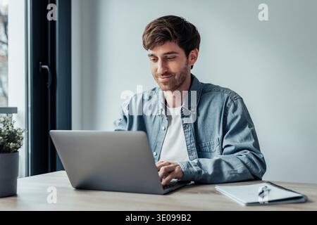 Side view handsome young businessman working with computer remotely, sitting at wooden table in office. Pleasant happy man communicating in social net Stock Photo