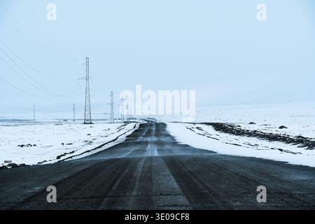 Desolate asphalt road stretches across snow covered Arabel Plateau with electric pylon line and gloomy sky in Kyrgyzstan Stock Photo