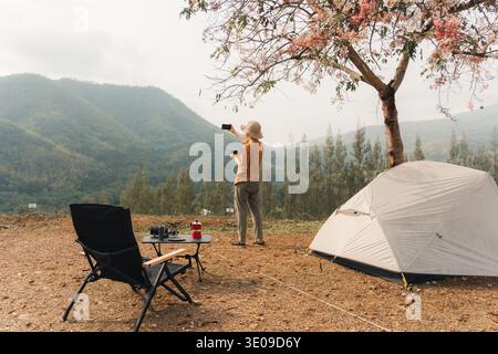 Solo traveler enjoying with using smartphone selfie at campsite with tent and camping equipment in mountain valley landscape during morning Stock Photo