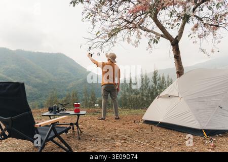 Solo traveler enjoying with using smartphone selfie at campsite with tent and camping equipment in mountain valley landscape during morning Stock Photo