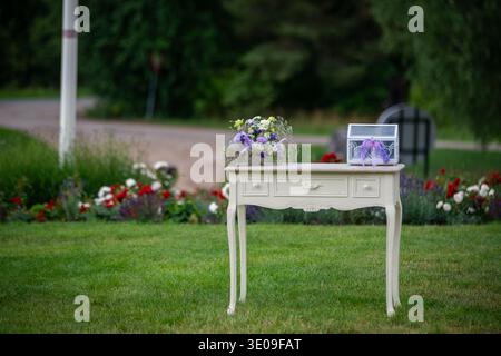 Elegant outdoor wedding setup featuring a vintage white table with a floral bouquet and decorative card box on green lawn. Romantic garden scene ideal Stock Photo