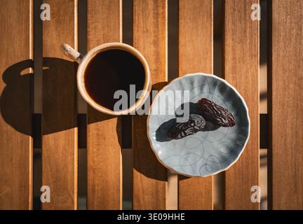 Top view of a cup of black coffee and two dried dates on a ceramic plate, arranged on a wooden table in warm morning sunlight for breakfast. Stock Photo