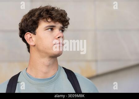 Teenage male standing by light stone wall on campus, wearing crewneck sweatshirt and backpack Stock Photo