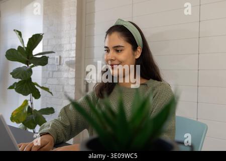 Woman working on laptop at home workspace, wearing green sweater and headband, blurred succulent Stock Photo