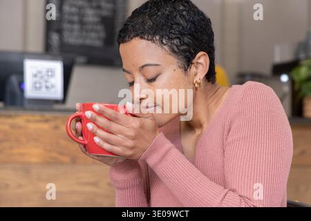 African American woman wearing pink ribbed sweater sitting in cafe holding red mug near QR code Stock Photo