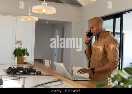 African American man standing at kitchen island using laptop and speaking on smartphone, copy space Stock Photo