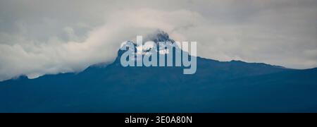 Moody landscape of a dark volcanic mountain peak with a small snow cap, wrapped in low clouds and mist under an overcast sky, panoramic composition. Stock Photo