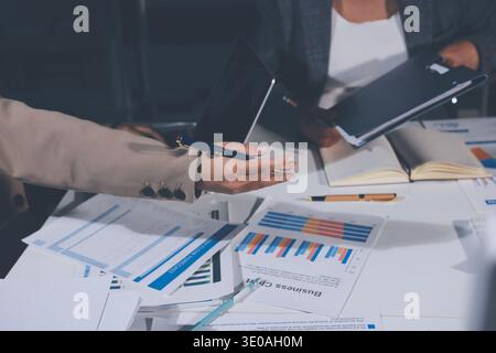 business documents on office table with tablet, smart phone and laptop and two colleagues discussing data Stock Photo