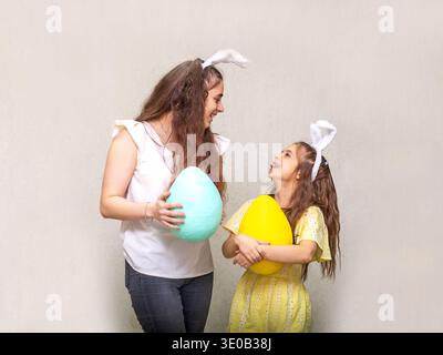Mother and daughter wearing bunny ears holding large pastel eggs and smiling during Easter celebration Stock Photo