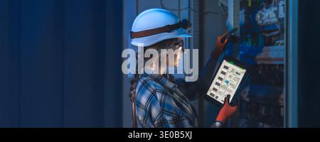 Female technician using tablet near electrical panel in blue light. Technician digital monitoring. Stock Photo