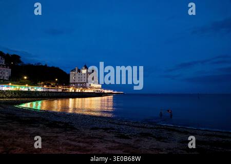 The coloured lights of the pier and Grand Hotel shimmer across a calm sea at Llandudno beach as evening brings dark shadows, North Wales. Stock Photo