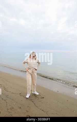 Young woman standing on sandy beach with arms raised toward calm sea. Freedom and peaceful coastal lifestyle moment. Stock Photo